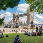 A Group Of People Sitting On Grass In Front Of A Bridge